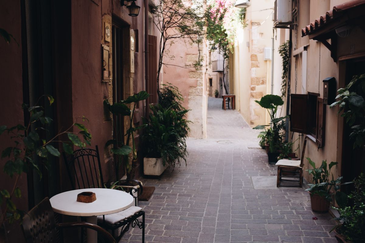 Narrow alleyway in Chania Old Town with flowering plants, stone walls, and outdoor café seating