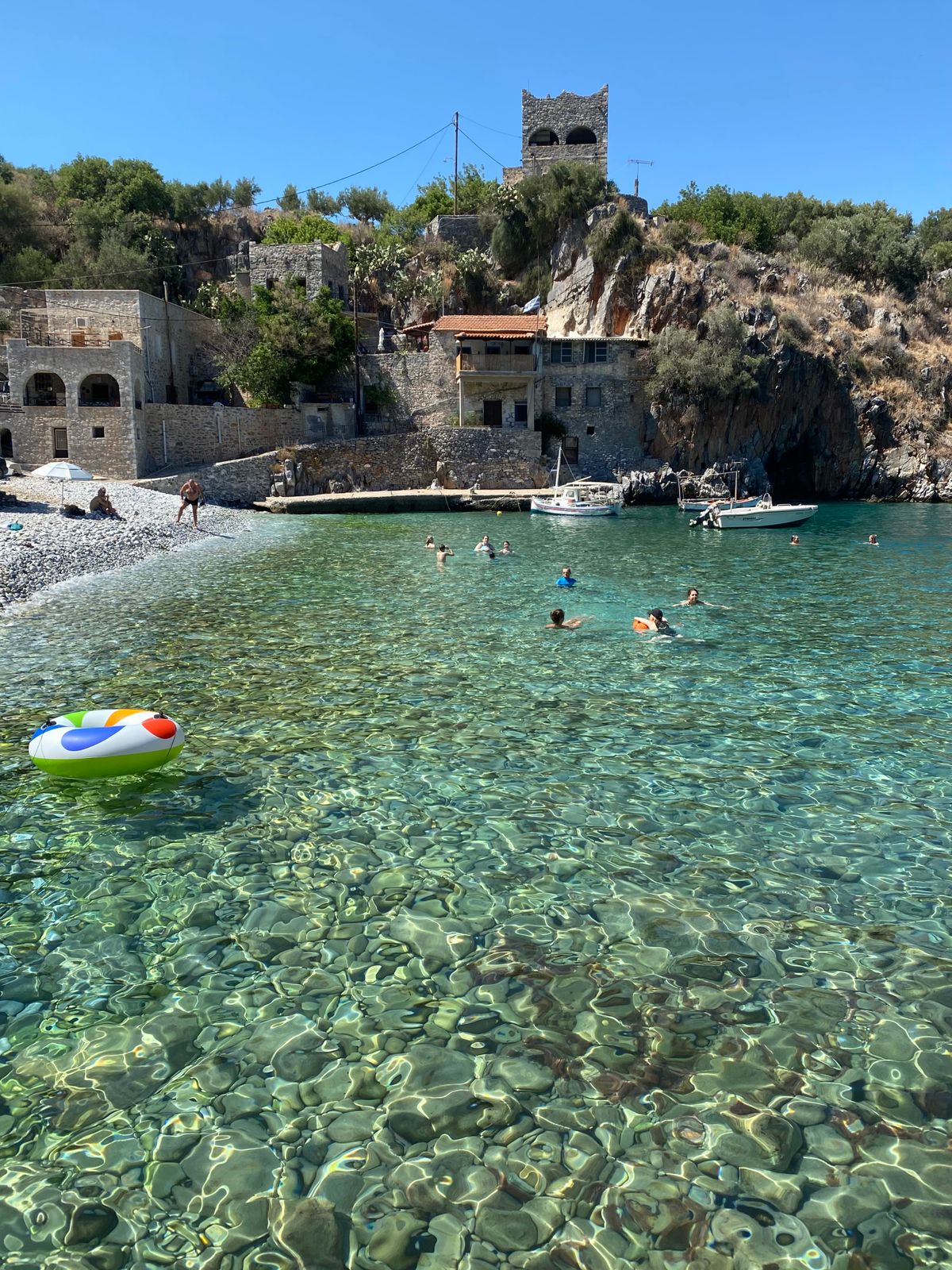 Swimmers in crystal clear waters beside stone architecture on the Mani Peninsula coast