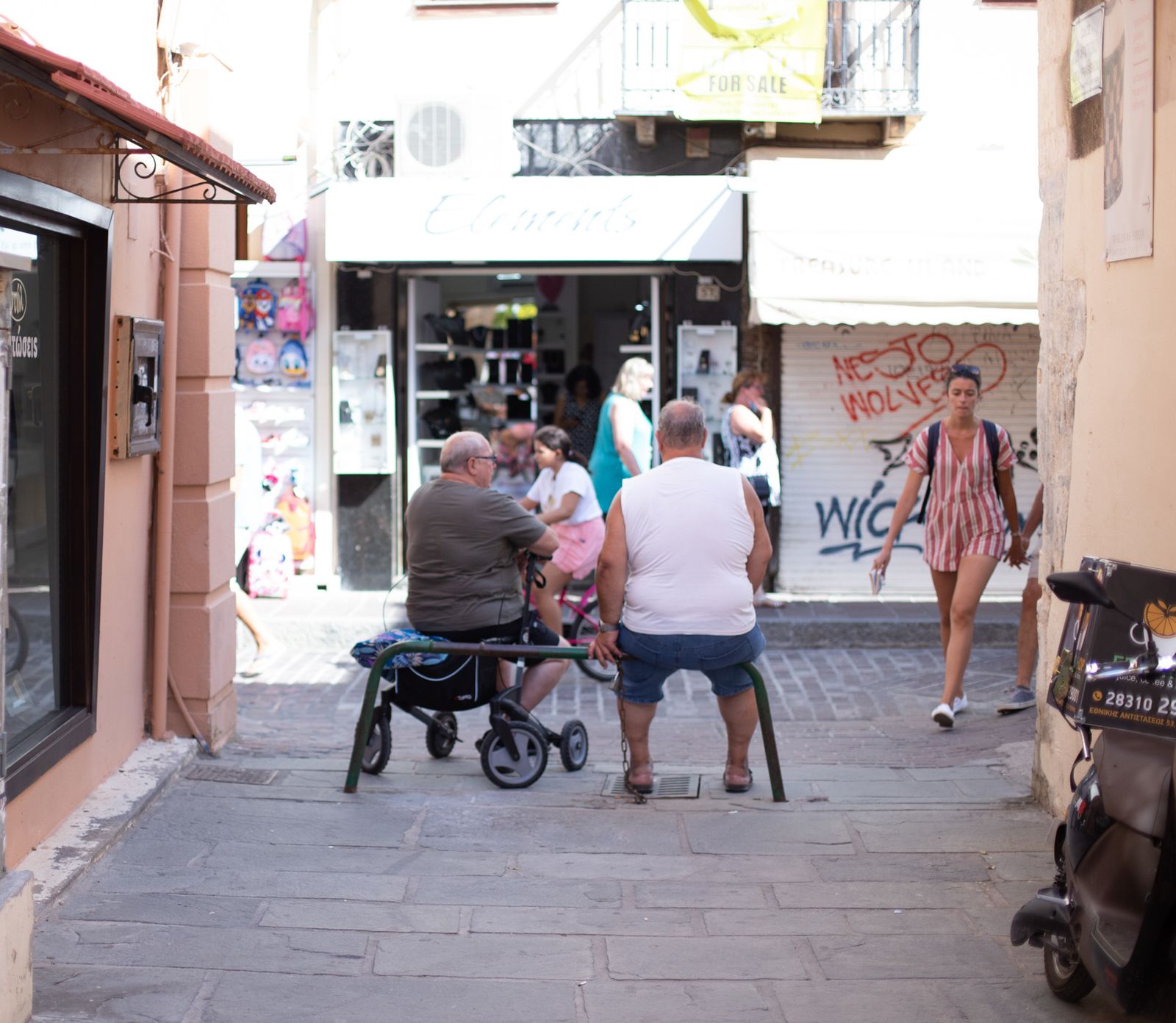 Pedestrians walking through a shaded cobblestone street lined with shops and cafes in Rethymno Old Town