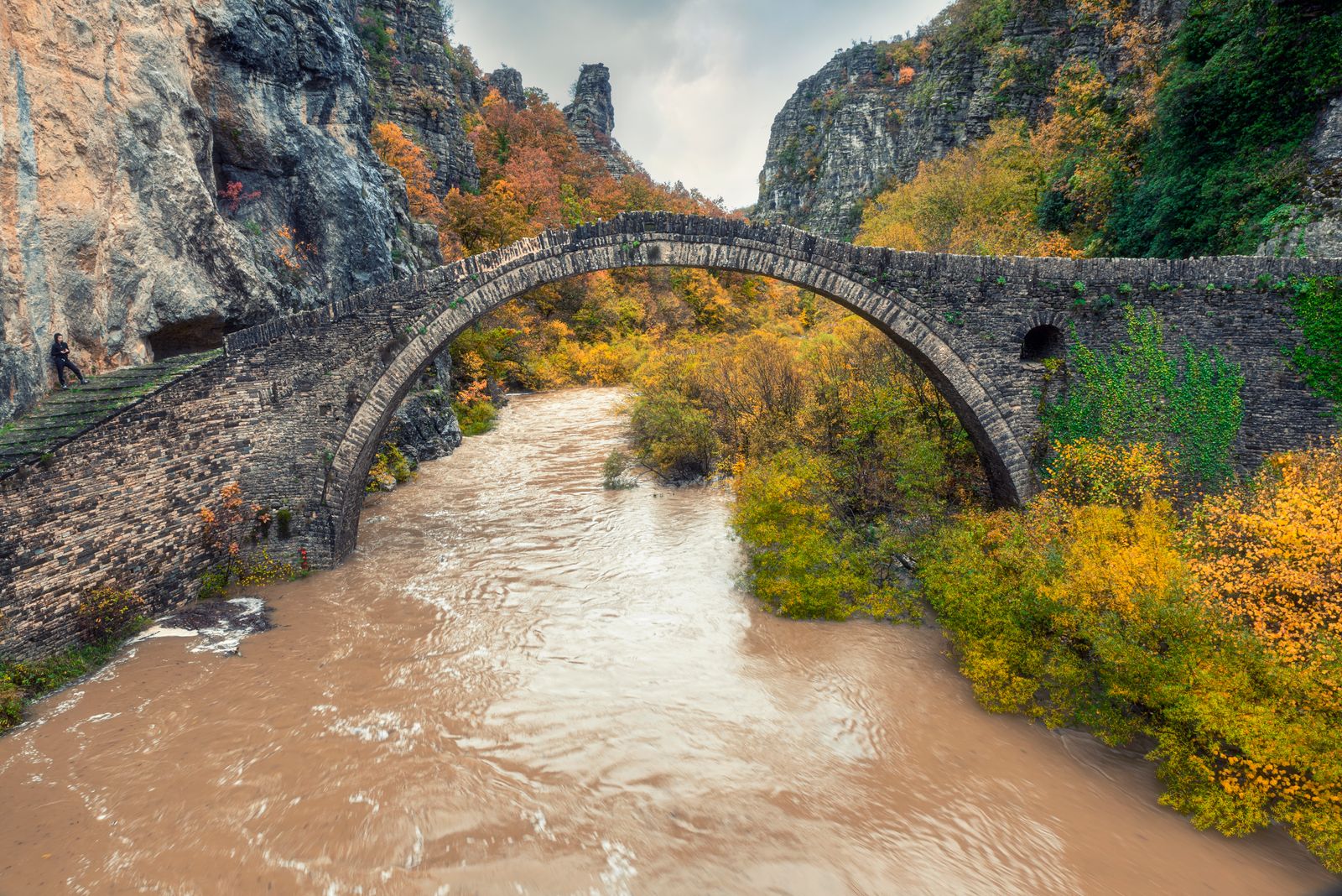 A single-arch stone bridge over a river in the Pindus Mountains during autumn, with golden foliage